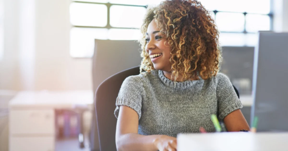 Smiling woman working at a desk with a laptop in a bright office setting.